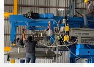 Two workers are operating and adjusting a large, blue industrial machine, while a third worker oversees the process from above wearing a yellow hard hat. The machine is labeled "Davis-Standard.