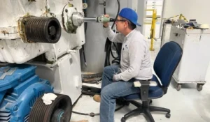 A person wearing a blue cap and safety glasses operates machinery while seated on a swivel chair in an industrial setting.