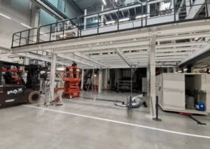 Industrial workspace with equipment, including a forklift, scissor lift, and various machinery. Elevated platform with railings above ground level adds additional working space.
