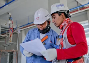 Two construction workers wearing safety gear and helmets review a document together inside a building under construction.