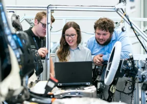 Three people, two men and one woman, working together on a laptop among car components in a laboratory setting.