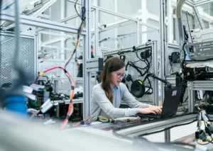 A woman in a high-tech lab works on a laptop surrounded by complex wiring and electronic equipment.