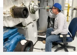 A person in protective eyewear and a blue cap is seated on a chair, operating a mechanical tool on a piece of industrial equipment.