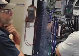 Two men, one with a beard and glasses, inspect an open electrical control panel with various wires, meters, and components visible.