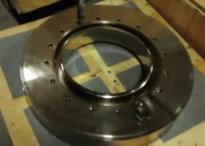 A large metallic circular part with numerous evenly spaced holes around its perimeter, placed on a padded surface.