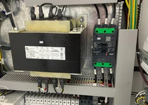 Electrical control panel featuring a transformer with labeled wiring and circuit breakers. Various cables and connectors are organized inside the enclosure.