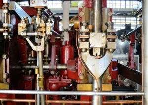 Industrial machinery with complex metal parts, gears, and mechanisms arranged in a dense assembly. Red and metallic components are prominent, indicating a heavy-duty mechanical apparatus.