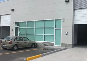 A beige car is parked outside a modern commercial building with glass windows and doors. The building’s exterior is light gray and marked with the numbers 52 and 60.