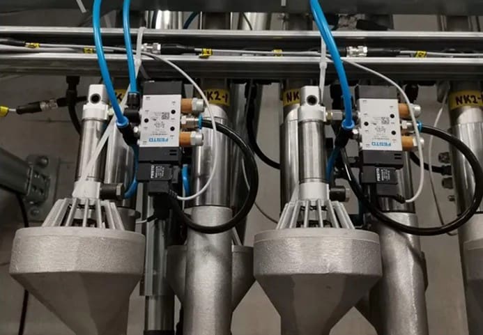 Close-up of an industrial machinery setup featuring metallic cylindrical components connected by blue and black tubes, with electronic regulators attached at the top.