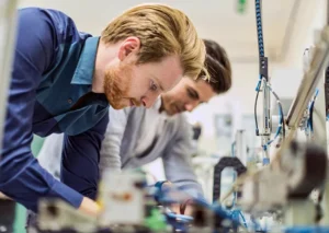 Two men are working in a laboratory, closely examining and handling equipment on a workbench.
