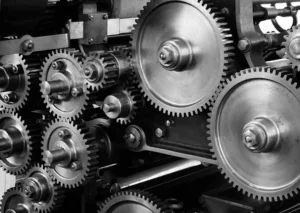 Close-up image of interlocking gears of various sizes in a mechanical assembly, demonstrating intricate engineering and machinery design. The gears are metallic and precisely aligned.