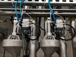 Image of industrial equipment with multiple metal components, hoses, and electrical connections. Several labels and wires are visible, indicating a setup for machinery or manufacturing processes.