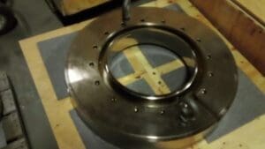 A metal ring with multiple holes sits on a wooden surface, partially placed on a foam mat.