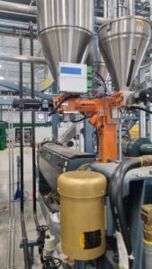 A manufacturing facility with industrial equipment, including metal containers, pipes, an orange robotic arm, and various control boxes.