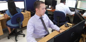 Three office workers are seated at desks using computers. The man in the foreground is focused on his screen, while a woman and another man are working in the background.