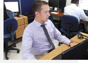 A man in a white shirt and tie is sitting at a desk working on a computer in an office with other people.