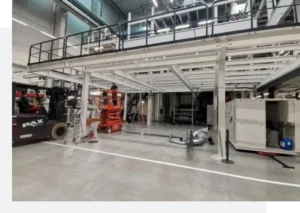 The image shows the interior of a large industrial facility, featuring scaffolding, machinery, and various equipment. A worker on a lift is present, and storage units are visible on the right side.